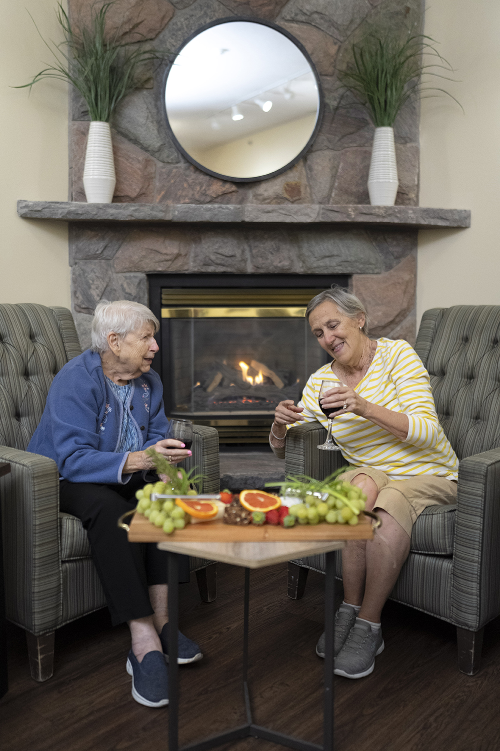 Two seated residents enjoying wine and cheese by the fireplace.