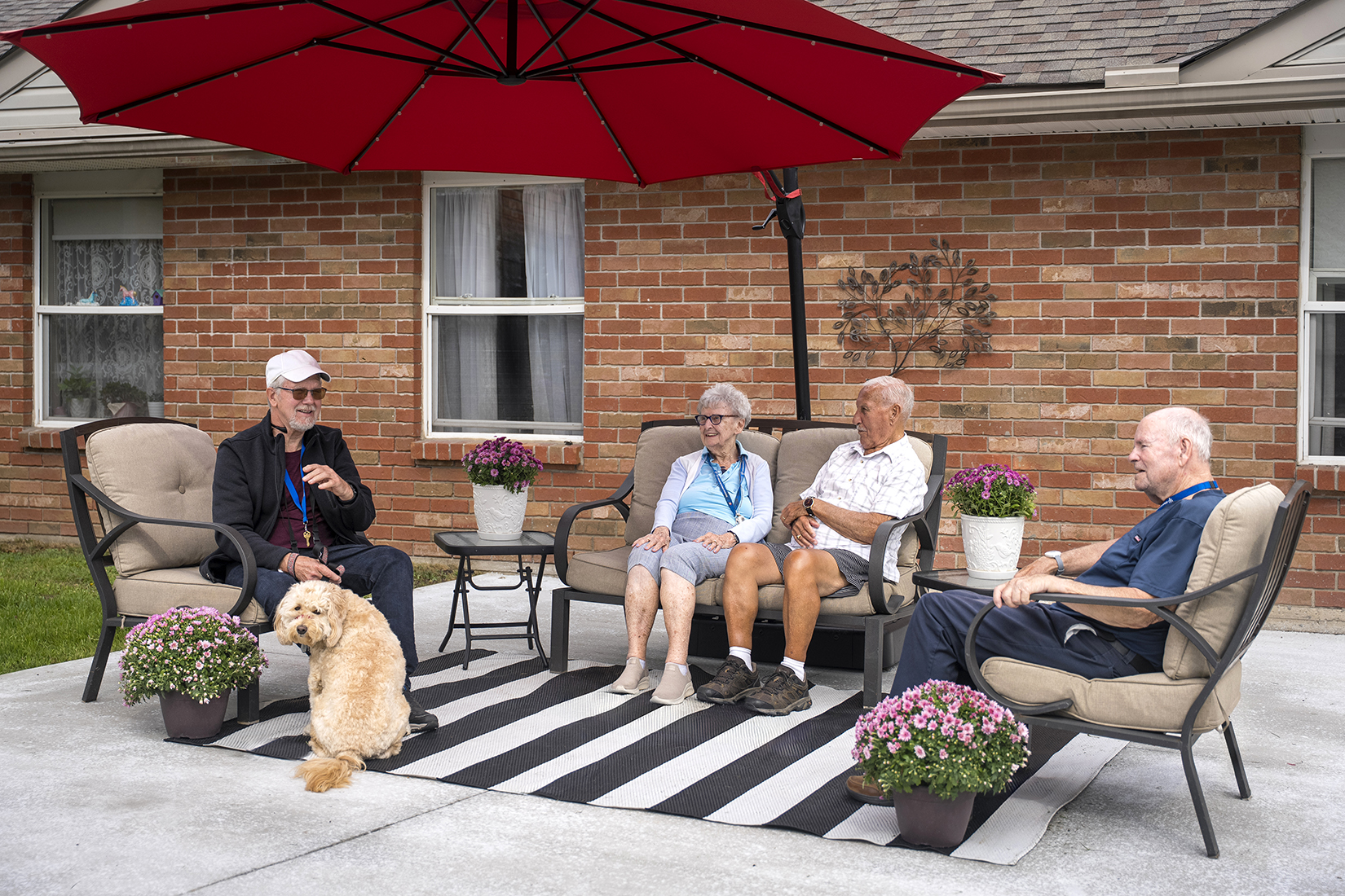 Residents relaxing with a dog under patio umbrellas on a warm fall day.